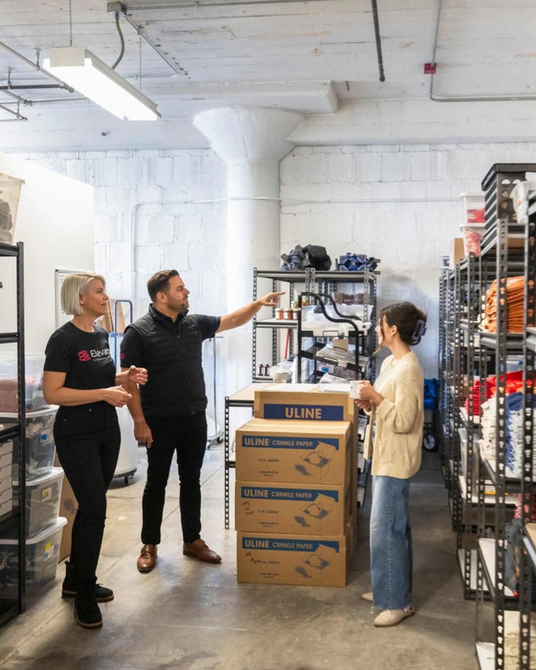 Elevator staff members talking with a business owner in a warehouse unit filled with shelving, storage bins, and packing supplies