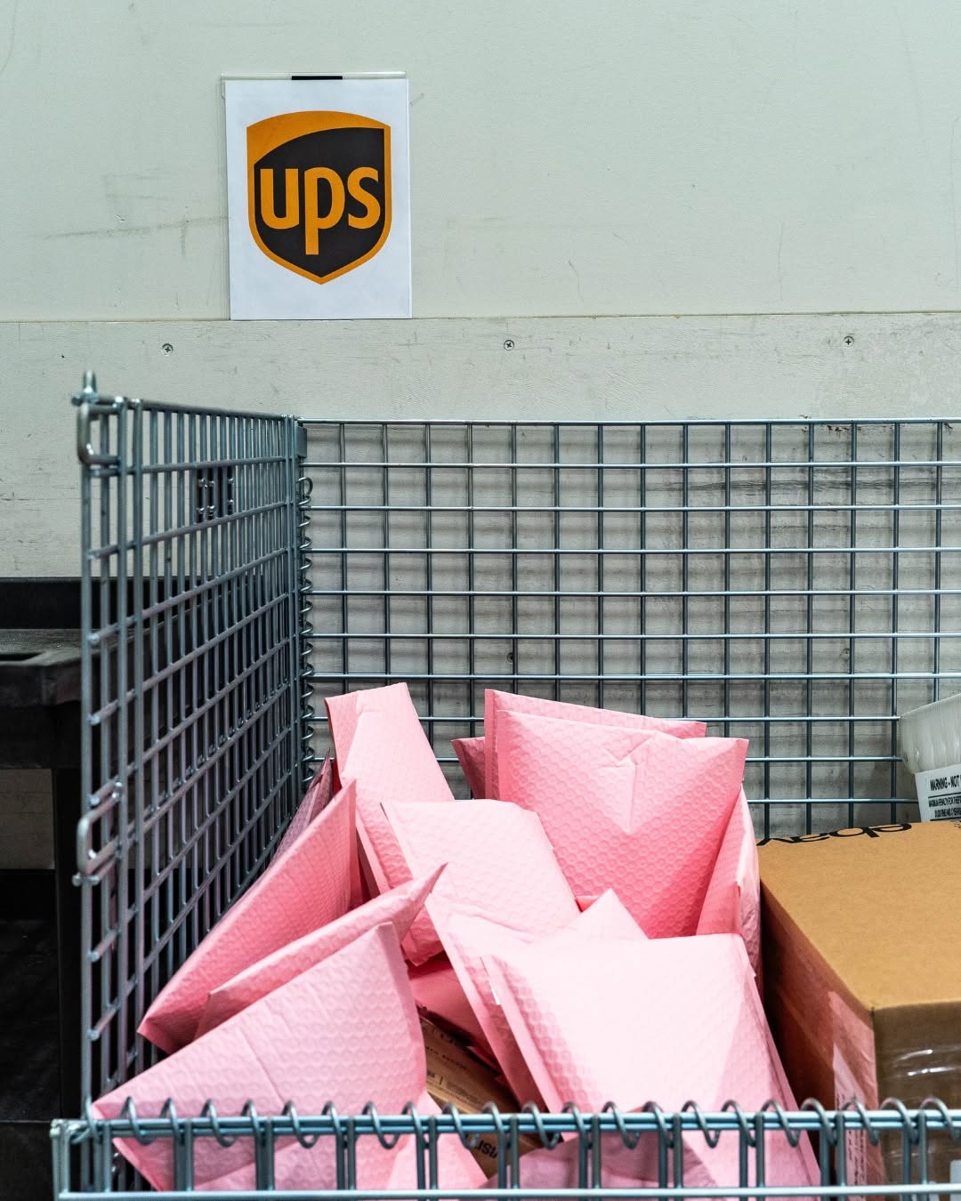 Wire cart holding pink bubble mailers and a shipping box staged beneath a UPS pickup sign at Elevator's Des Moines location
